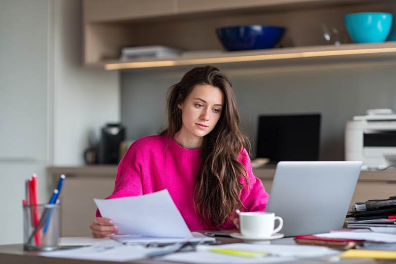 A young woman (18-19 years old) with long dark hair working at a desk in a modern office, wearing a pink sweater, holding a piece of paper in one hand and typing on a laptop with the other, a laptop with a blank screen, a white coffee cup with a saucer, a container with colored pens and pencils, scattered papers and notebooks on the desk, an office printer and light-colored cabinets in the background, a blue bowl on a shelf, clean and organized workspace, natural lighting, highly detailed, realistic, 16:9 aspect ratio --ar 3:2 --v 7 Job ID: 7c1abe01-9fab-492e-b110-aa9e6ff5f8a6